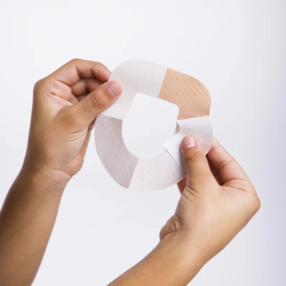 Two hands holding the omnipod tan adhesive patch and peeling off the backing paper on one of the 4 sections