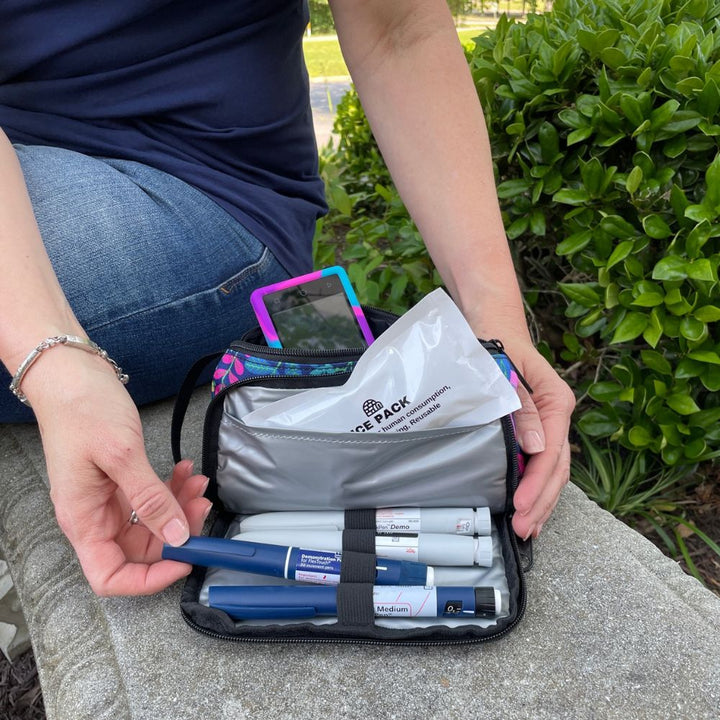 A women sitting with the blue floral pattern Insulated Diabetes Insulin Supply Case open on her lap pulling an insulin pen out of secured loop.