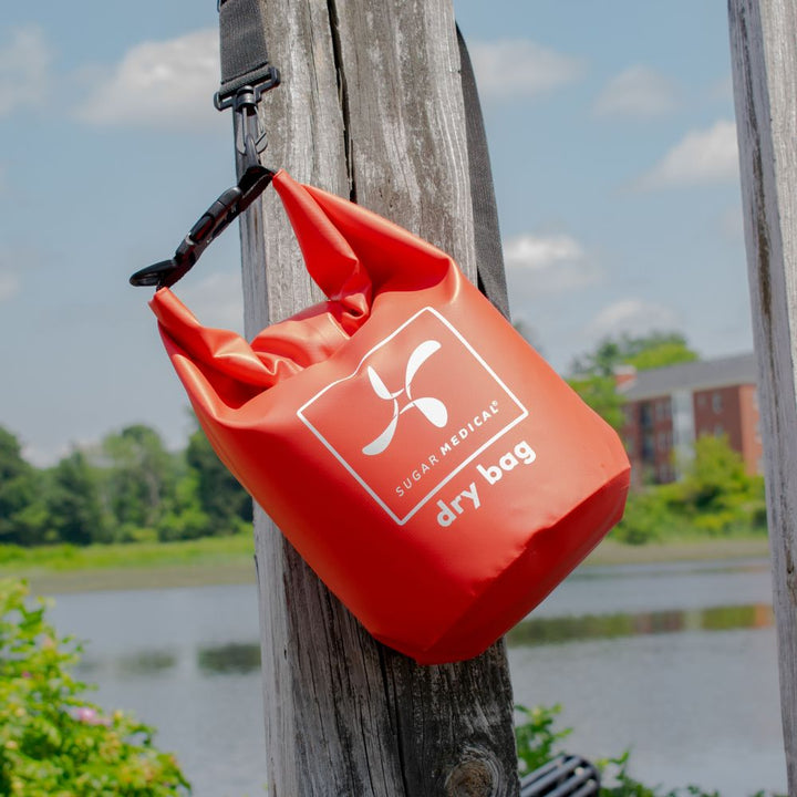 Sugar Medical red dry bag hooked with shoulder strap on fence in front of water.