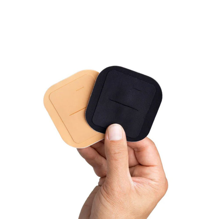 Hands holding two Skin Grip adhesive patches for the Tandem Mobi insulin pump — one in black and one in tan.