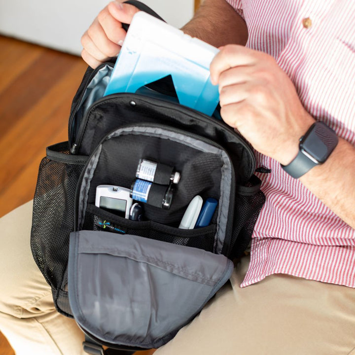 Diabetes Insulated Sling Backpack in black opened with diabetic supplies organized in it on men’s lap pulling out ice pack from insulates compartment.