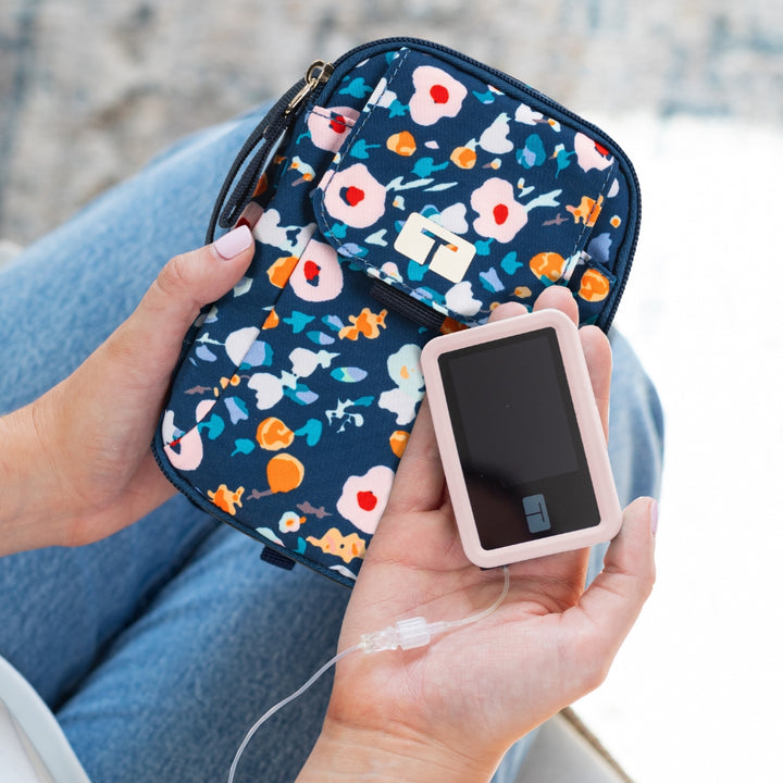 Women's right hand holding Tandem Insulin pump with a light pink silicone gel skin on and a blue floral Tandem supply case in other hand,