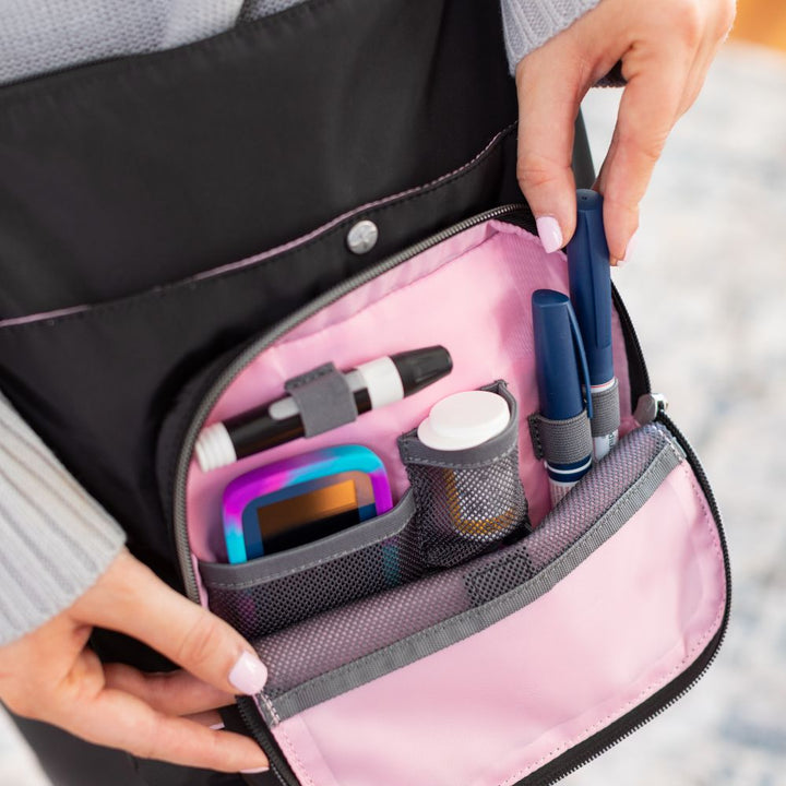 Diabetes Nylon Backpack in black front compartment set up with glucose meter, test strips, insulin vial, lancet and insulin vial worn on women’s shoulder.