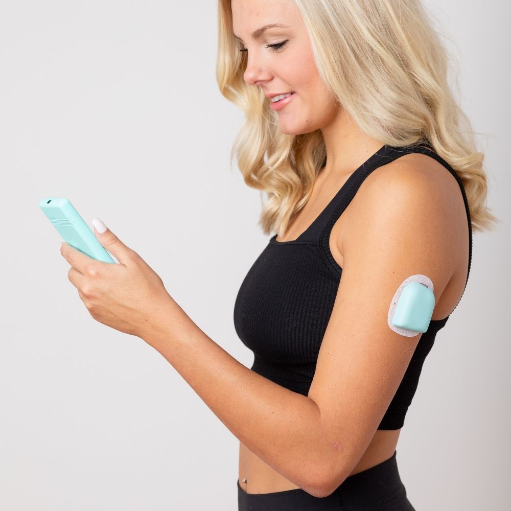A woman looking at her omnipod device while wearing the aqua PumpPOP on her upper left arm