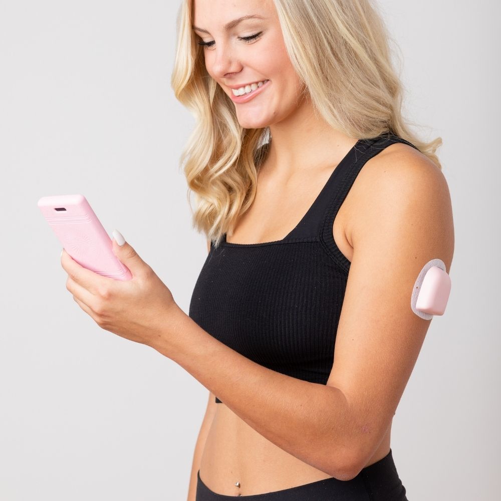 A woman looking at her omnipod device while wearing the light pink PumpPOP on her upper left arm