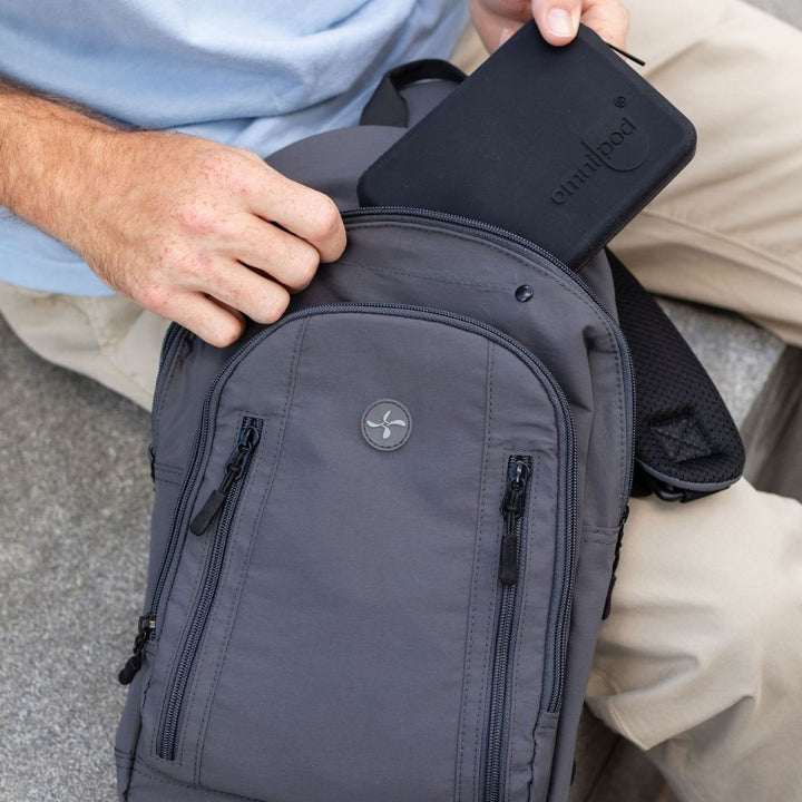 A close-up of a dark gray backpack being worn, with a person's hands placing a black rectangular device into the top pocket. The backpack has multiple zippered compartments and a circular logo on the front.