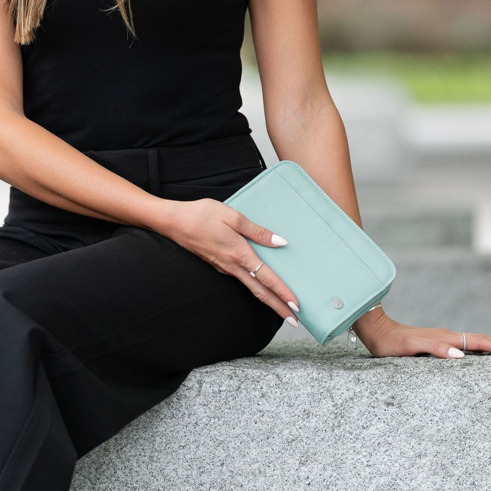 A woman in a black outfit sits on a stone ledge holding a seafoam diabetes supply case with a silver snap button.