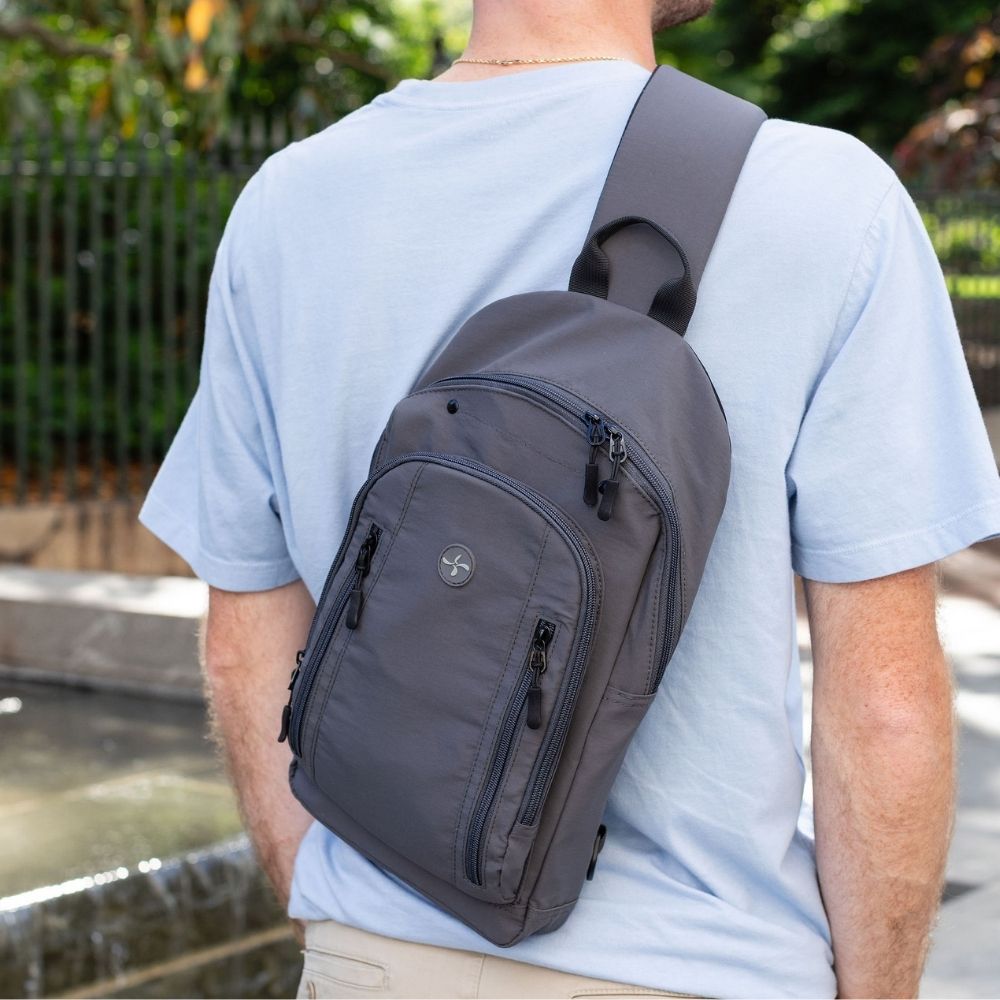 A man wearing a light blue t-shirt and khaki pants, viewed from behind, wearing a dark gray crossbody sling backpack with multiple zippered pockets.