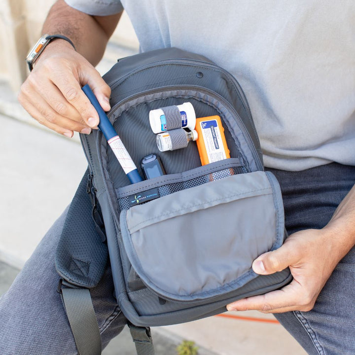 Man pulling out a insulin pen from the front pocket of the charcoal grey Insulated Roam Sling Backpack with diabetic supplies organized. 