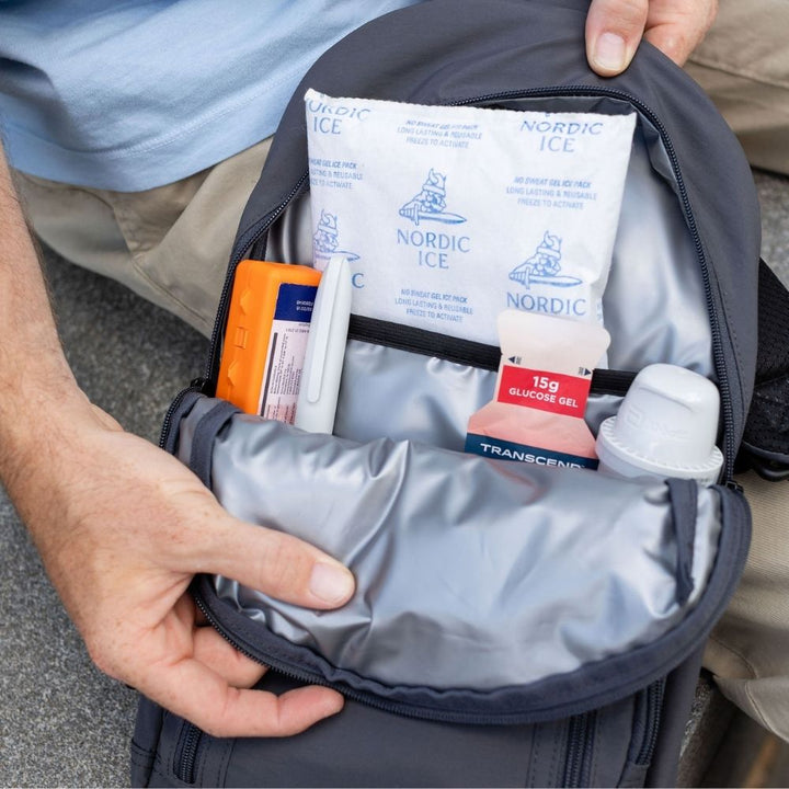 An open gray backpack shown from above, containing Nordic Ice gel packs and other small medical or travel items, with a person's hands holding the bag open.