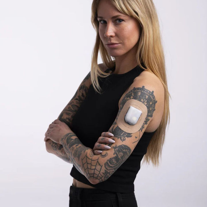 A woman with her arms crossed in a black tank top wearing the tan omnipod adhesive patch around the pod on her upper right arm