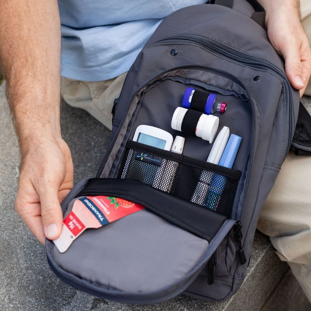 A close-up of a gray backpack being held open by a person, showing the front pocket filled with medical and personal items such as a blood pressure monitor, pens, and a card holder.