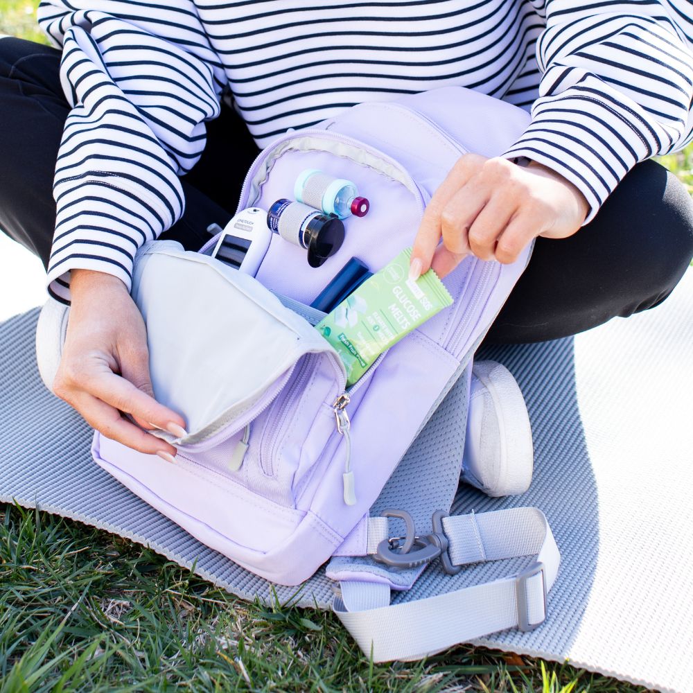 Girl opening the front pocket of a light purple Insulated Roam Sling Backpack organized with diabetic supplies pulling out glucose. 