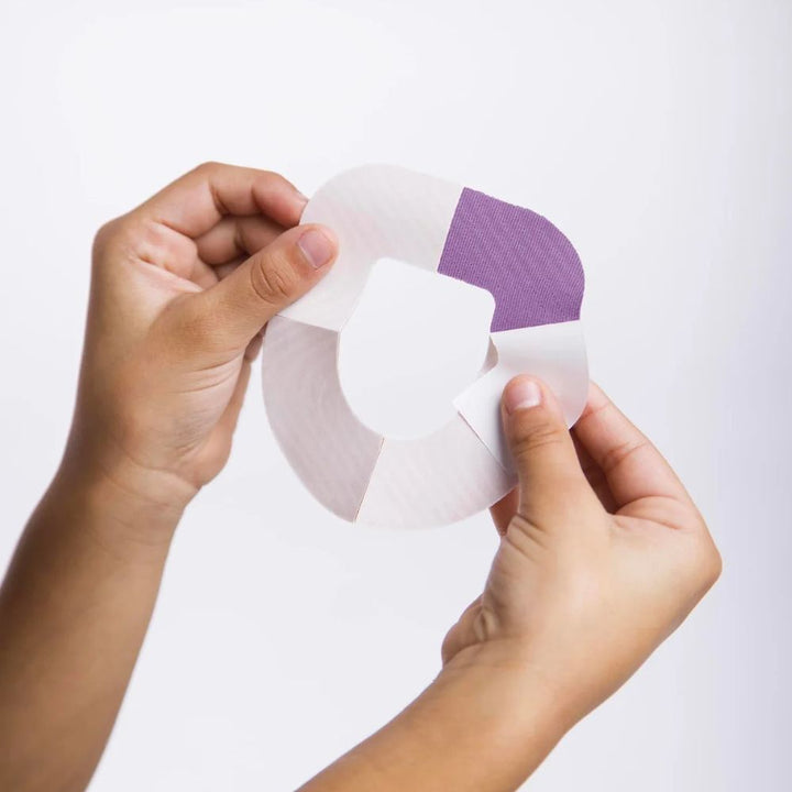 Two hands holding the omnipod purple adhesive patch and peeling off the backing paper on one of the 4 sections 