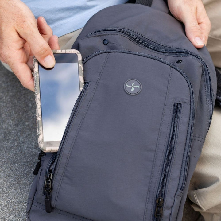 A close-up of a dark gray backpack being held by a person, with a smartphone inserted into a front slip pocket. The person's hands and legs are visible, along with a light blue shirt and khaki pants.