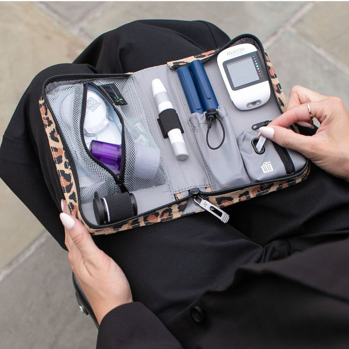 A leopard-print carrying case opened on a person's lap, holding a blood glucose meter, lancets, a pen-style device, and other small medical accessories. A hand is visible holding the case open.