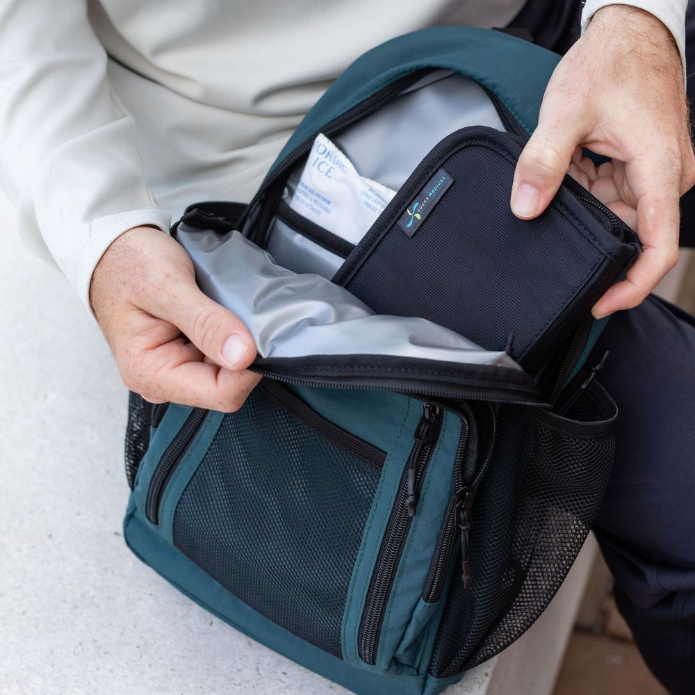 A close-up of a person's hands opening a dark teal and black diabetes backpack to reveal its interior compartments, including a zippered pocket and a light gray lining.