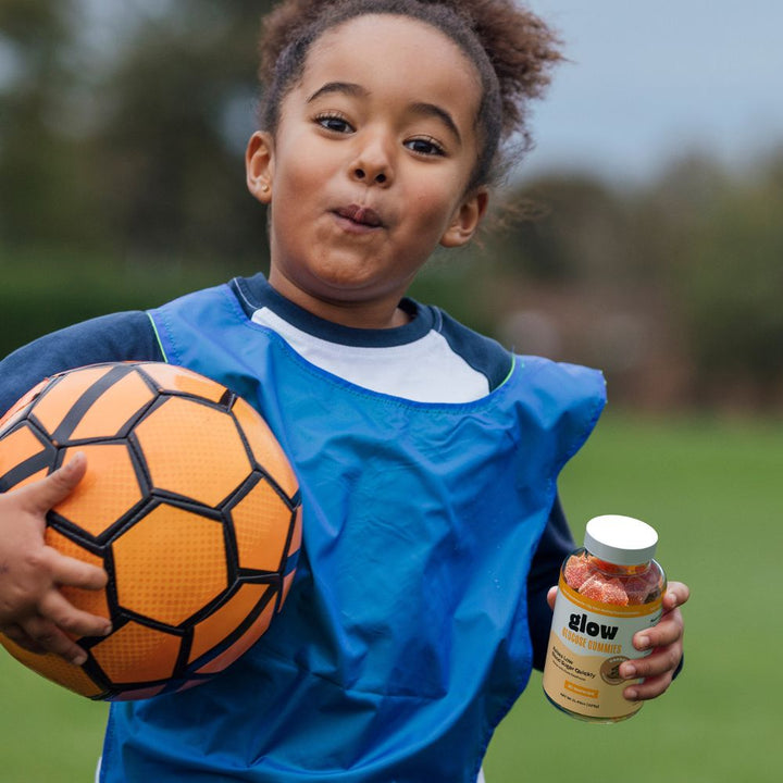 A child holding a soccer ball in one hand and a glow glucose gummies bottle in the other hand. 