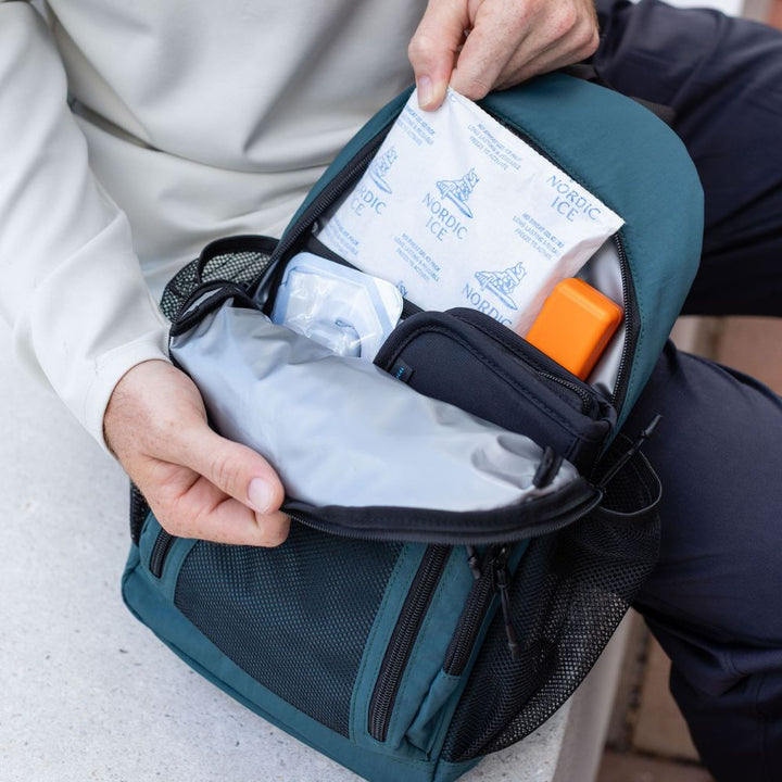 A close-up of a teal and black diabetes backpack being opened by a person, showing contents including a packet of Nordic Ice gel, a glucagon, and a diabetes supply bag.