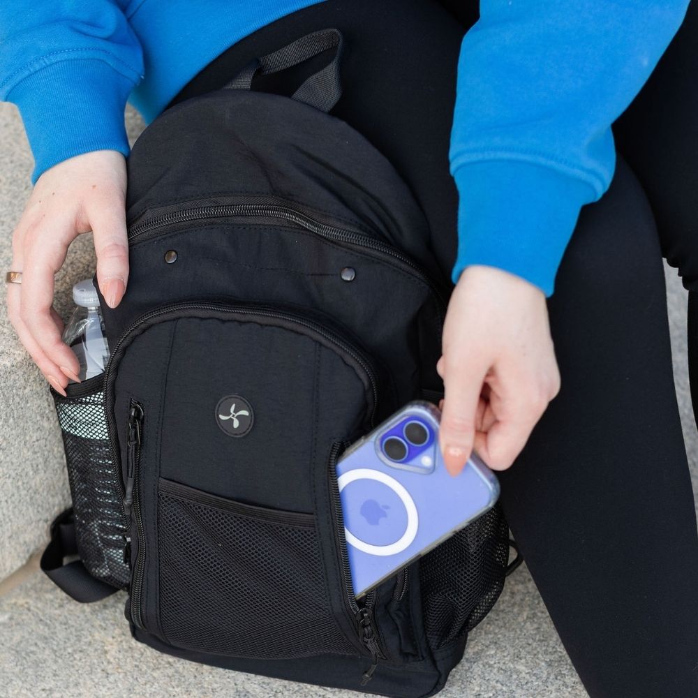 A black backpack with a side mesh pocket and zippered front compartment, shown being used while seated. A person in a bright blue sweatshirt is placing a blue smartphone into the pocket.