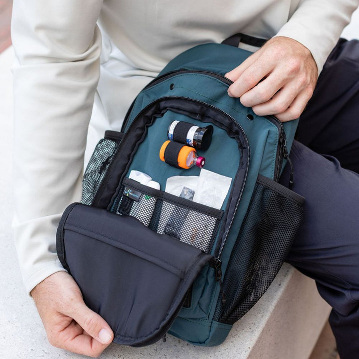 A close-up of a teal and black medical-style backpack being opened by a person, showing mesh pockets with diabetes supplies. The person is wearing a light beige long-sleeve shirt and dark pants, seated on a light stone surface.