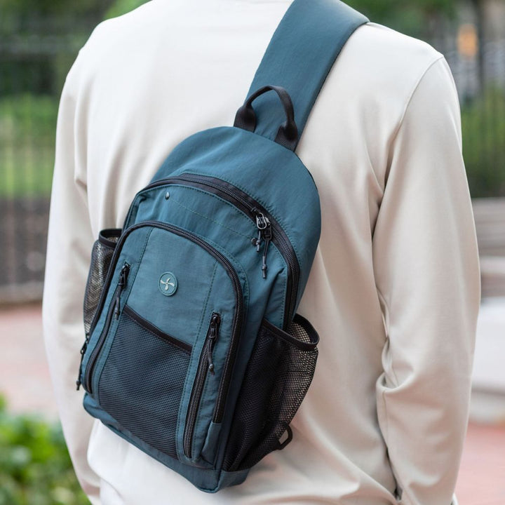 A person wearing a light beige long-sleeve shirt shown from the back, wearing a teal blue sling backpack with black mesh side pockets and black zippers.