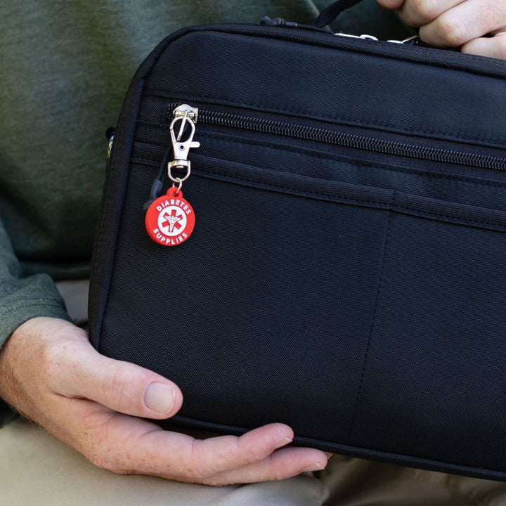 Person holding a black travel bag with a red diabetes supply charm attached to the front zipper.