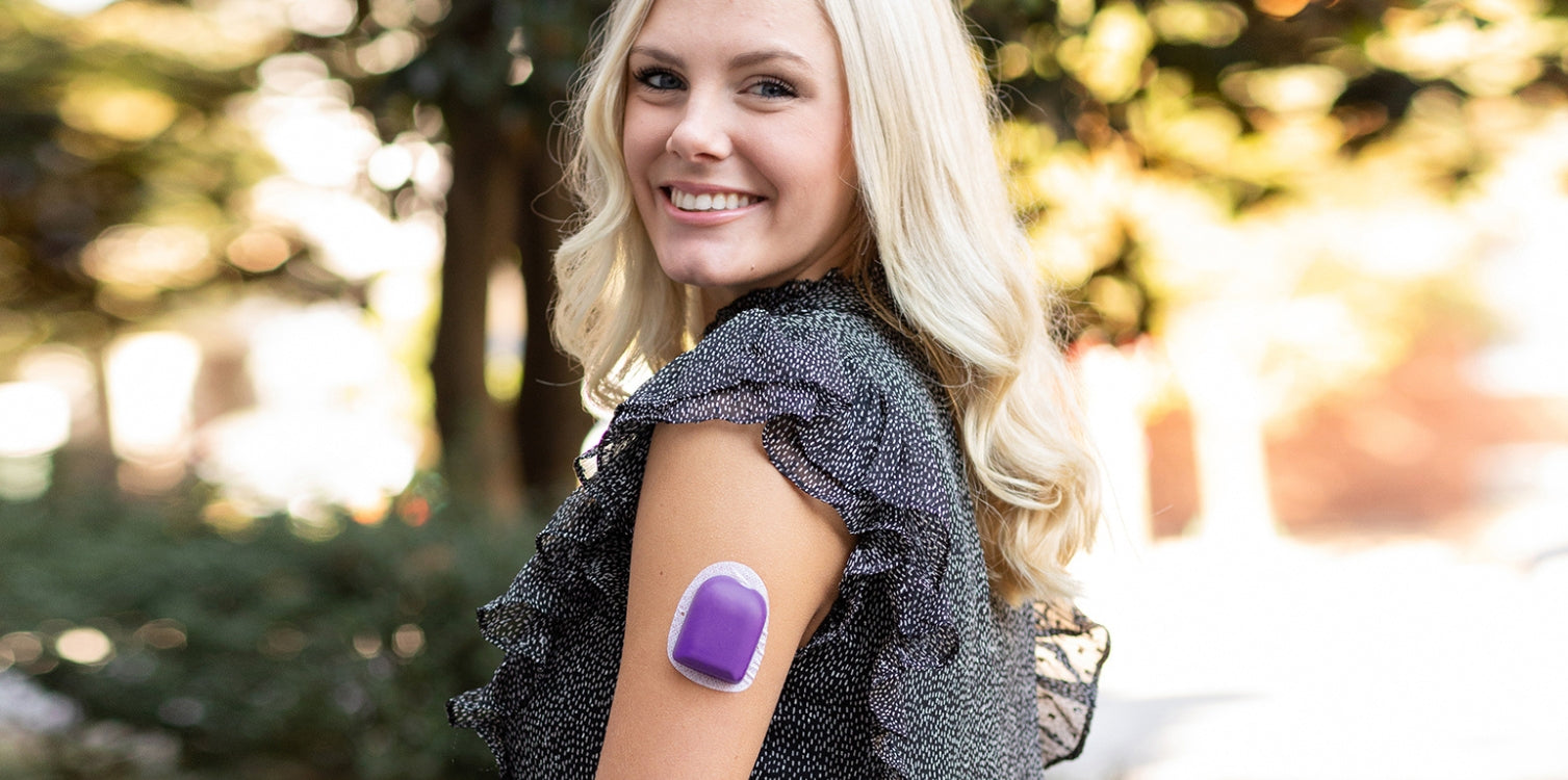 Smiling woman outdoors wearing a black patterned blouse with ruffled sleeves, showing a purple insulin pump cover on her upper arm.