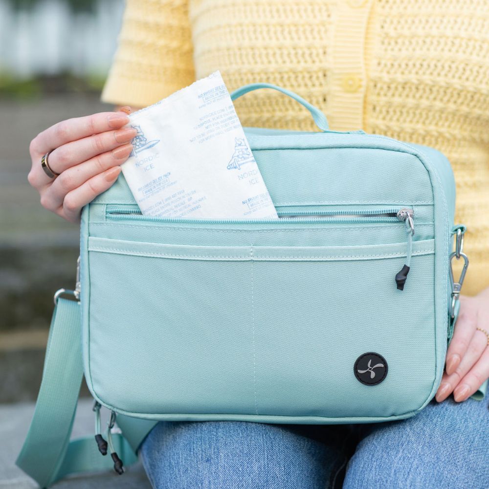 A seafoam crossbody bag with a top handle and front zip pocket, shown being held by a person wearing a yellow knit sweater and blue jeans. A ice pack is tucked into the top zipper.