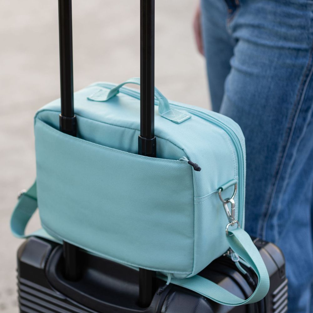 A seafoam travel diabetes bag with a top handle and side strap, shown clipped onto a black rolling suitcase with a person in blue jeans standing nearby.