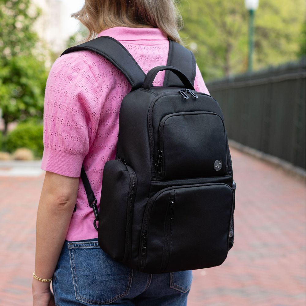 Person wearing a black backpack outdoors on a brick path with greenery.