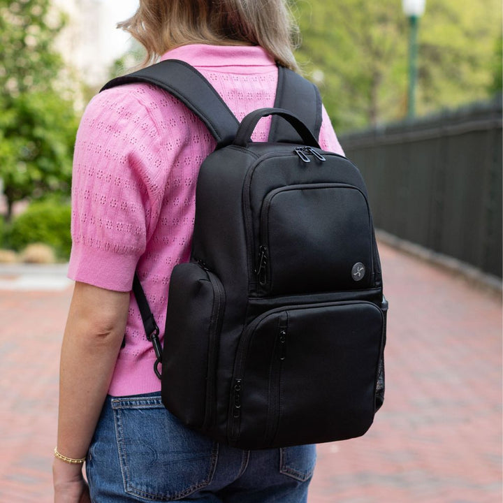 Person wearing a black backpack outdoors on a brick path with greenery.