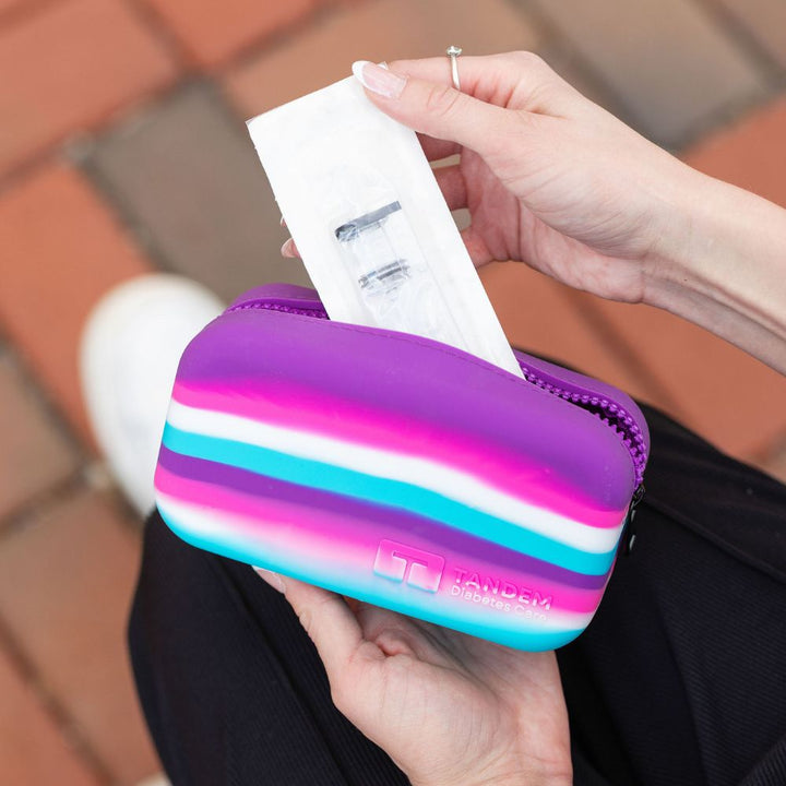 Person holding a colorful case with Tandem diabetes supplies, on a blurred outdoor background.
