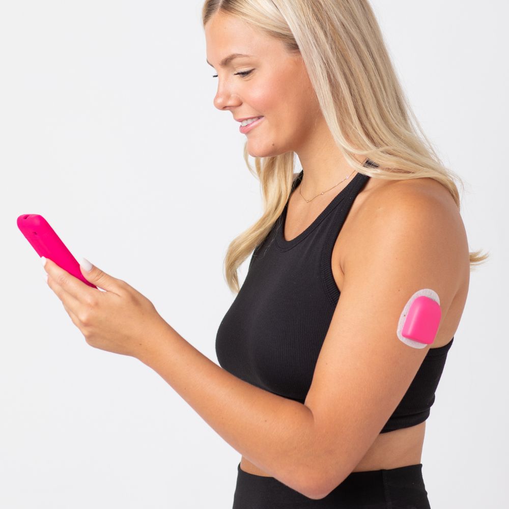 A woman looking at her omnipod device while wearing the raspberry (bright pink) PumpPOP on her upper left arm