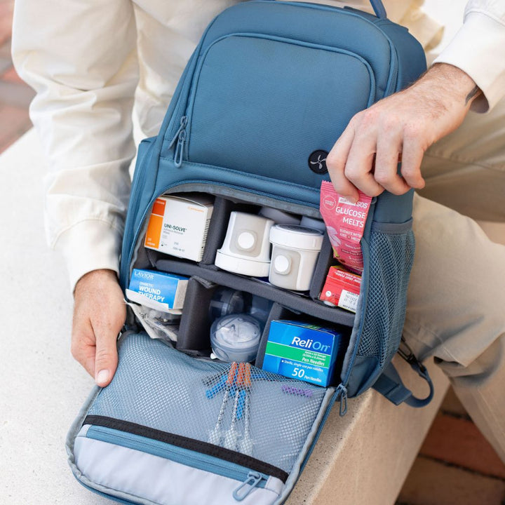 A person wearing a cream shirt and khaki pants sits beside an open Steel Blue medical-style backpack, showing organized compartments filled with first-aid items such as bandages, gauze, and boxed supplies.