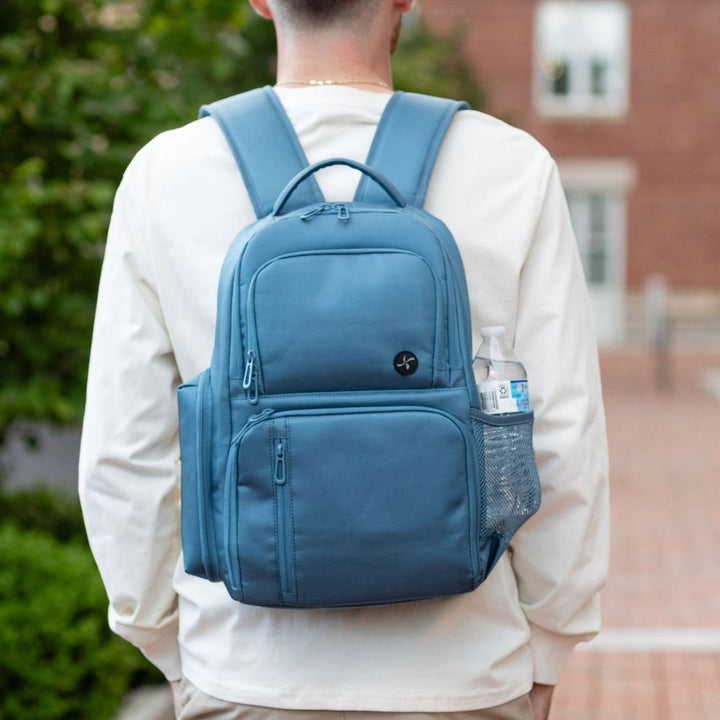 Person wearing a Steel Blue backpack outdoors on a brick path with greenery.