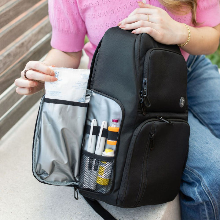 Person sitting on a bench with an open insulated pocket on side of black backpack containing ice pack and insulin.