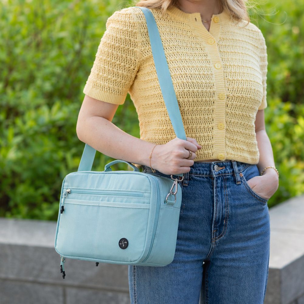 A woman wearing a yellow knit short-sleeve top and blue jeans carries a seafoam diabetes travel bag with a matching strap, photographed outdoors against a blurred green hedge.