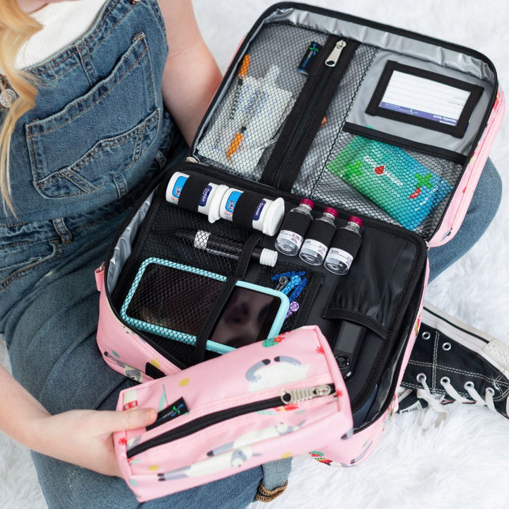 Diabetes Insulated Travel Bag in pink with llamas back compartment for a glucagon kit, Tandem supplies, Dexcom, snacks, glucose tablets, and includes a removable pouch on girl’s lap. 